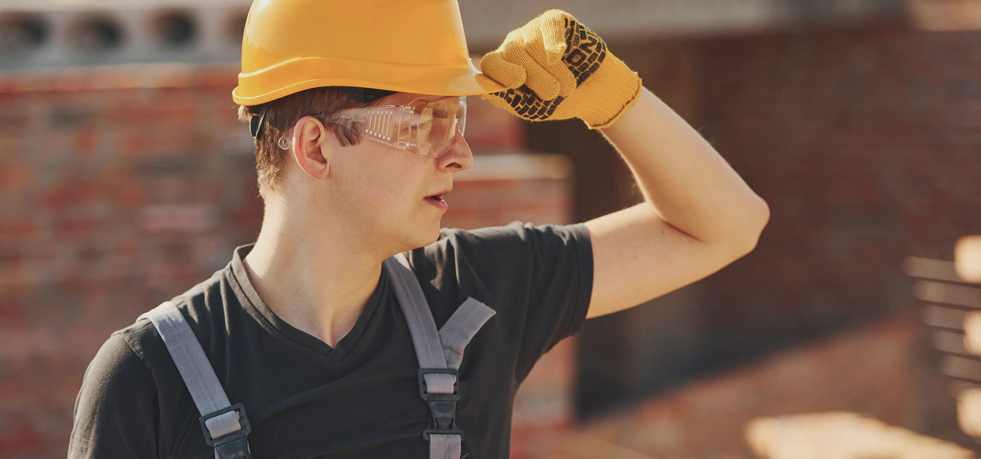 Osoba w kasku ochronnym i rękawicach roboczych stoi na placu budowy, osłaniając dłonią oczy przed światłem